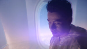 Alejandro Londoño en una escena del videoclip LEJOS observando por la ventana de un avión en una atmósfera cinematográfica con iluminación púrpura.